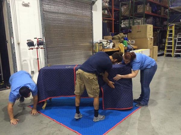 Three movers in a warehouse wrapping a large table in a quilted blue moving blanket. The table sits on a blue floor pad, with warehouse shelving and boxes in the background.